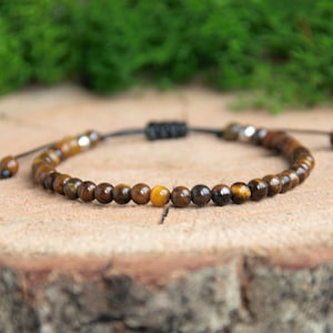 Könnte beinhalten: Ein braunes und goldenes Perlenarmband mit schwarzem Band und silbernen Akzenten. Das Armband besteht aus Tigeraugen-Steinen.