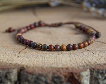 May include: A beaded bracelet featuring small, round, multi-coloured natural stone beads in shades of brown, red, and ochre. The bracelet is strung on a thin, dark brown cord with an adjustable sliding knot closure at the back for a secure fit.