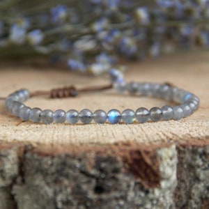Può includere: Un braccialetto in pietra di labradorite blu con chiusura in corda marrone. Il braccialetto è fatto di piccole perle rotonde e viene mostrato su una superficie di legno.