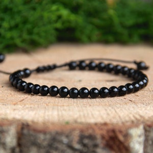 Könnte beinhalten: Ein schwarzes Perlenarmband mit einem einfachen, verstellbaren Kordelverschluss. Das Armband besteht aus kleinen, runden Perlen und wird auf einer Holzoberfläche gezeigt.