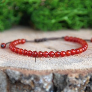 Può includere: Un braccialetto di perline rosse con chiusura a nodo scorrevole. Il braccialetto è composto da piccole perline rotonde e lucide. Le perline sono di un colore rosso vibrante. Il braccialetto è esposto su una superficie di legno con muschio verde sullo sfondo.