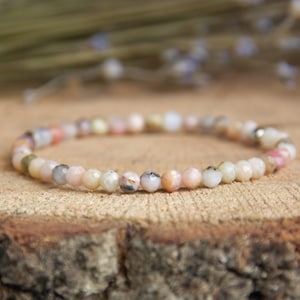 Peut inclure: Un bracelet de perles délicat avec de petites pierres précieuses rondes multicolores, roses, crème et grises. Le bracelet est monté sur un élastique et repose sur une surface en bois rustique, pour un style naturel.