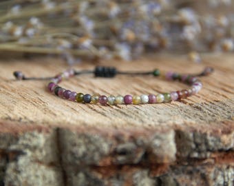 May include: A delicate bracelet with small, multi-coloured beads in pink, green, yellow, and brown. The beads are strung on a black cord with an adjustable knot. The bracelet is displayed on a wooden surface.