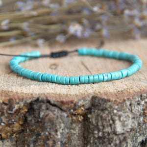 Op de afbeelding: Een turquoise kralenarmband met een verstelbaar zwart koord. De armband is gemaakt van kleine, cilindrische kralen en wordt gepresenteerd op een rustiek houten oppervlak. De achtergrond bevat gedroogde planten.