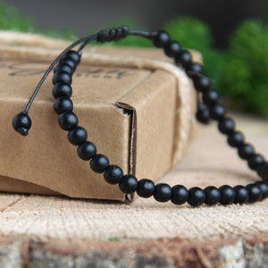 May include: A black beaded bracelet with a black cord and adjustable closure. The bracelet is made of small, round beads and is lying on a wooden surface.