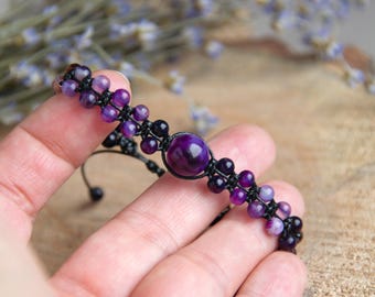 Puede incluir: Una pulsera de macramé hecha a mano con cuentas de amatista en tonos morados y una piedra central grande de color morado oscuro. La pulsera se sostiene en una mano, con un fondo borroso de lavanda seca y madera.