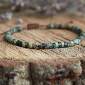 Puede incluir: Una pulsera de cuentas con pequeñas cuentas cilíndricas en tonos verdes, azules y marrones. La pulsera está ensartada en un cordón marrón con un cierre de nudo corredizo ajustable. La pulsera se exhibe sobre una superficie de madera.