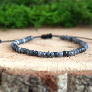 May include: A gray gemstone bracelet with black cord and a sliding knot closure. The bracelet is made with faceted beads and has a subtle ombre effect.
