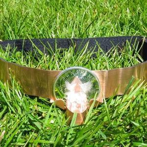 May include: A gold-colored headband with a large, clear crystal in the center. The crystal is surrounded by a silver-colored metal frame with a design that resembles a sunburst.
