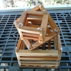 May include: Two wooden orchid baskets stacked on top of each other. The baskets are square with a slatted design and metal hardware. The wood has a natural, light brown color. The baskets are sitting on a black, grid-like surface.