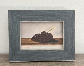 May include: A framed painting of a cluster of dark purple grapes with a green stem. The artwork is set against a light background and a brown surface. The frame is a muted gray color. The artist's signature is in the lower left corner.