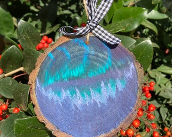 May include: A round wooden ornament with a painted aurora borealis scene in shades of blue and green. The ornament is trimmed with a natural bark edge and has a black and white checkered ribbon for hanging.