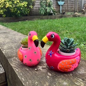 May include: Two pink flamingo planter pots with green and orange accents. Each pot has a small succulent plant inside.