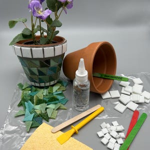 May include: A mosaic flower pot with purple flowers, a terracotta pot, and a bottle of glue. Also included are mosaic tiles in shades of green and white, a yellow spreader, and wooden sticks. A craft project.