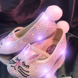 May include: A pair of pink sparkly baby shoes with a bunny design and white pom-poms. The shoes are covered in iridescent rhinestones and have a black outline of a bunny on the toe.