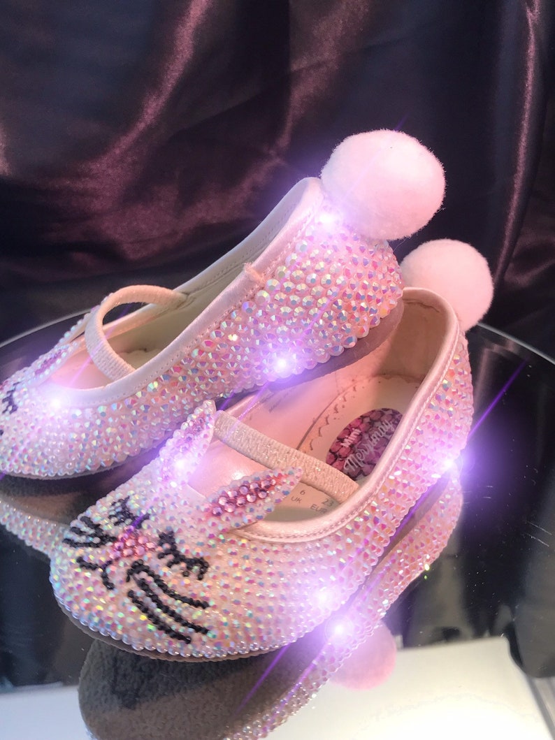 May include: A pair of pink sparkly baby shoes with a bunny design and white pom-poms. The shoes are covered in iridescent rhinestones and have a black outline of a bunny on the toe.