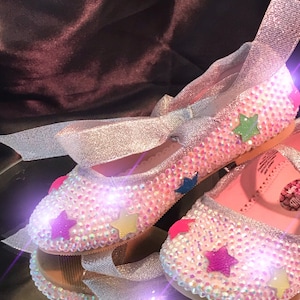 Puede incluir: Un par de zapatos plateados brillantes con un lazo y adornos de estrellas de colores. Los zapatos están decorados con pedrería iridiscente.