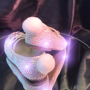 May include: A pair of white ballet flats with a sparkly iridescent finish and white pom-poms on the toes. The shoes are reflected in a mirror surface.