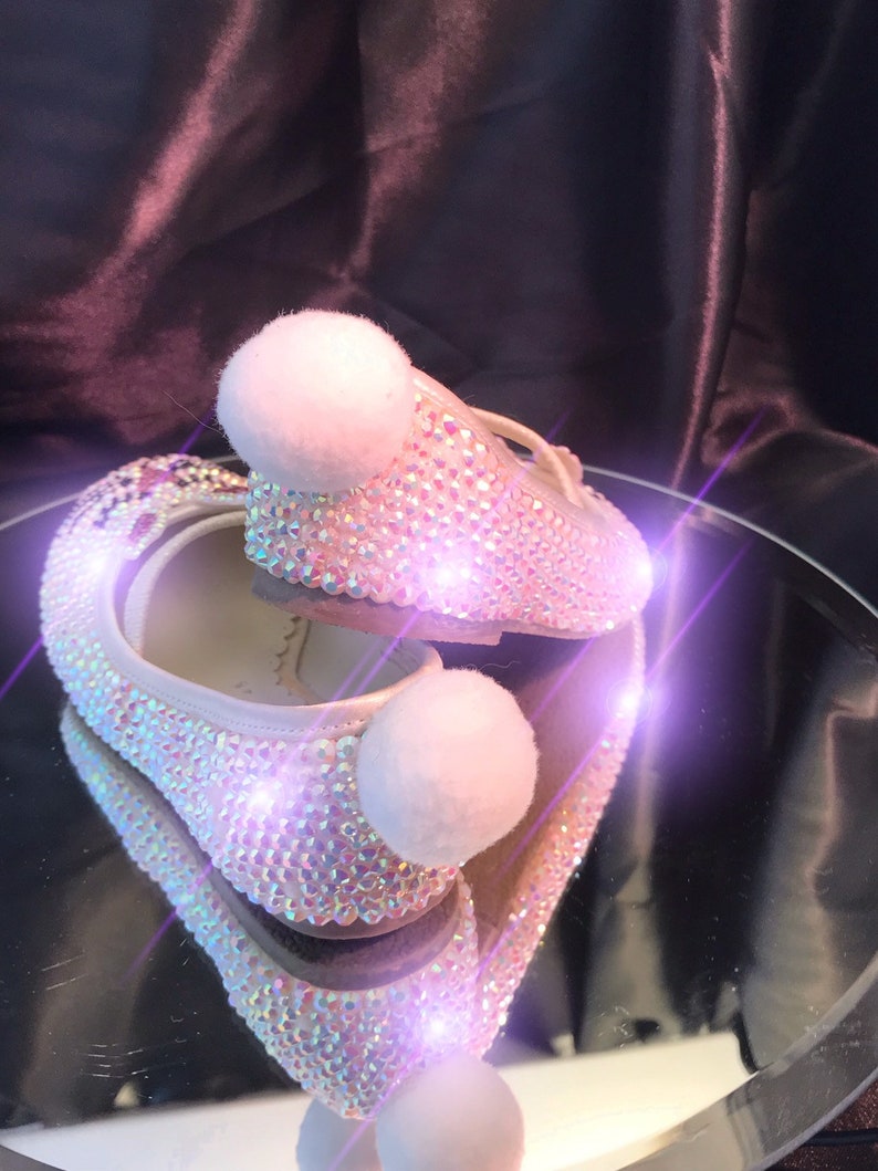 May include: A pair of white ballet flats with a sparkly iridescent finish and white pom-poms on the toes. The shoes are reflected in a mirror surface.