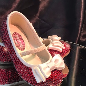 Könnte beinhalten: Ein Paar rote glitzernde Schuhe mit weißen Satinbändern. Die Schuhe haben einen weißen Riemen und sind mit roten Strasssteinen verziert. Die Schuhe sind in China hergestellt.