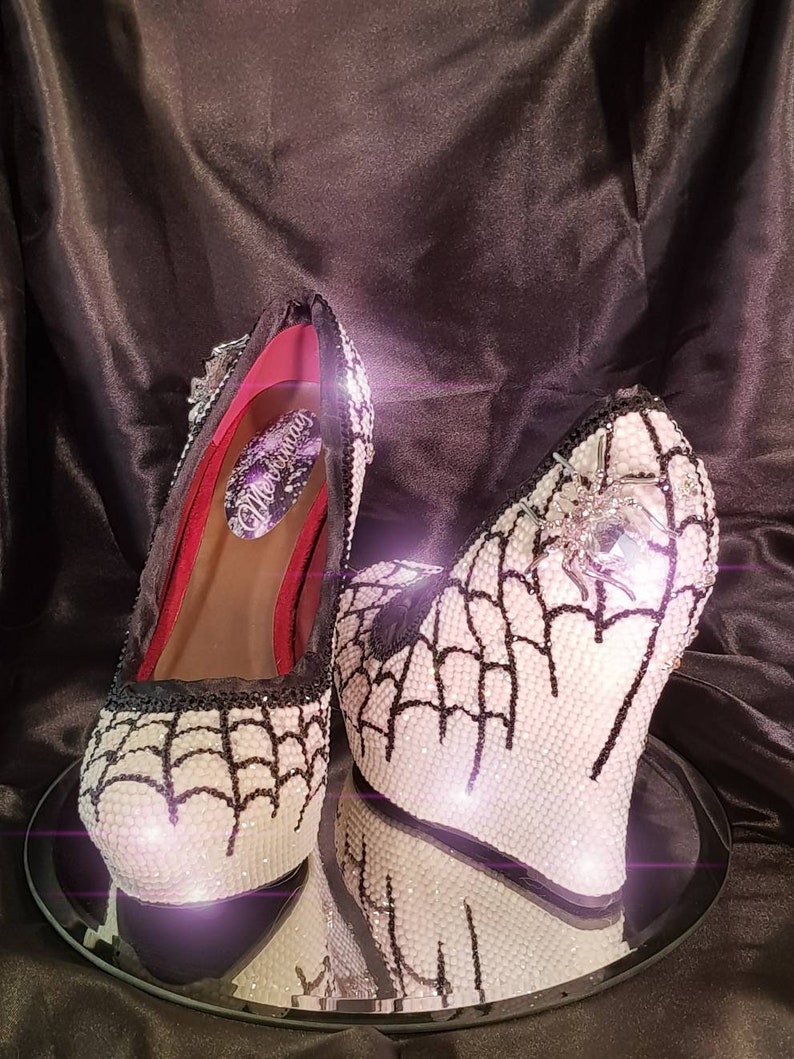 May include: White wedge heels with a black spiderweb design and a silver spider embellishment. The heels are covered in rhinestones.