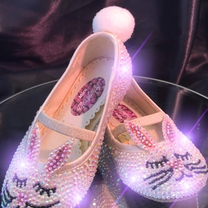 May include: A pair of white satin children's shoes with a bunny rabbit design. The shoes are covered in iridescent rhinestones and have a white pom-pom on the top. The shoes are lit up with pink and purple lights.