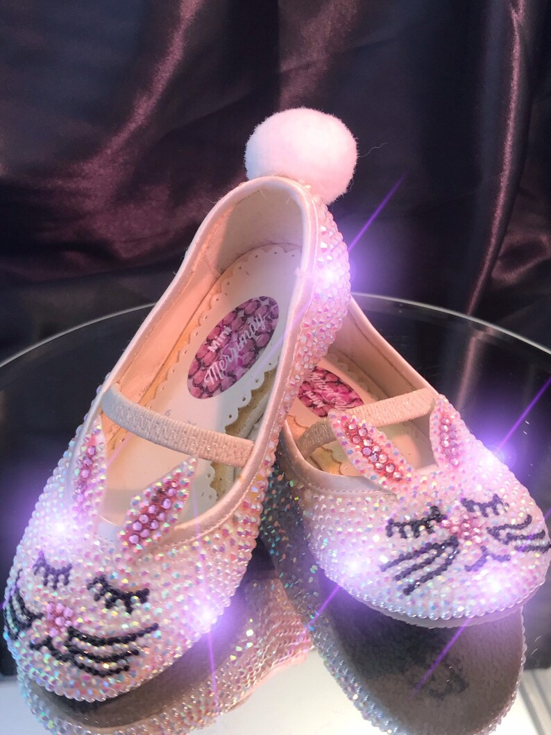 May include: A pair of white satin children's shoes with a bunny rabbit design. The shoes are covered in iridescent rhinestones and have a white pom-pom on the top. The shoes are lit up with pink and purple lights.