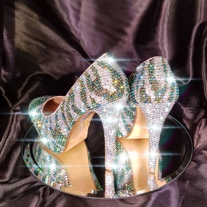 Könnte beinhalten: Ein Paar hochhackige Schuhe, die mit schimmernden grünen und klaren Strasssteinen besetzt sind und ein Zebra-Muster bilden. Die Schuhe stehen auf einer spiegelnden Oberfläche.