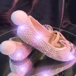 May include: A pair of pink sparkly shoes with fuzzy white pom-poms on the toes. The shoes are covered in iridescent rhinestones and have a small strap across the foot. The shoes are made in Vietnam.