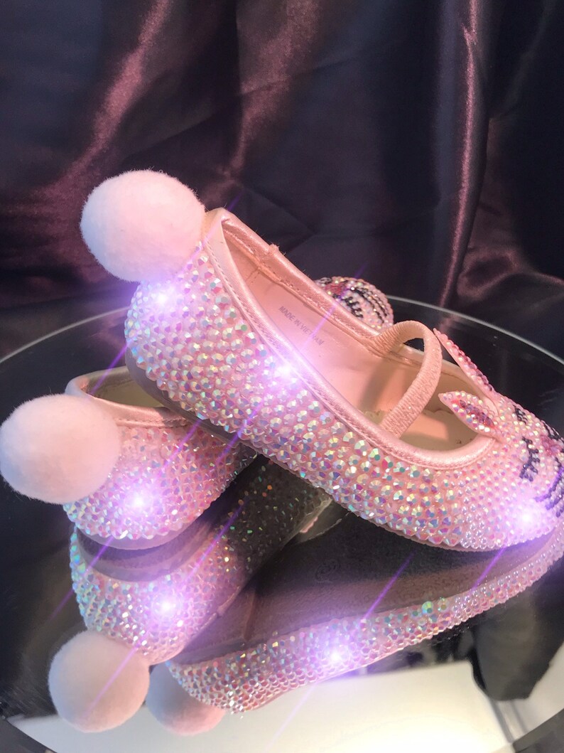 May include: A pair of pink sparkly shoes with fuzzy white pom-poms on the toes. The shoes are covered in iridescent rhinestones and have a small strap across the foot. The shoes are made in Vietnam.