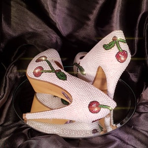 May include: A pair of white high-heeled shoes with a cherry design. The shoes are covered in rhinestones and have a red cherry with a green stem on each shoe.