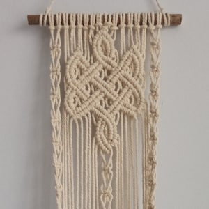 May include: A macrame wall hanging with a geometric design, made with natural cotton cord. The hanging is attached to a wooden dowel and has a plant in the bottom.
