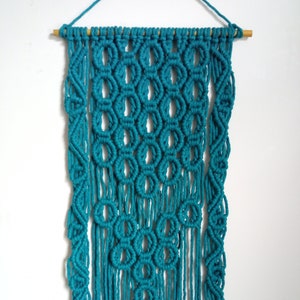 May include: Teal macrame wall hanging with a geometric pattern and fringe. The hanging is made with cotton cord and is attached to a wooden dowel.