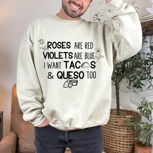 May include: A cream-colored crewneck sweatshirt featuring black text that reads: ROSES ARE RED VIOLETS ARE BLUE I WANT TACOS & QUESO TOO. The design includes small line-art illustrations of a rose, a flower, a taco, and a bowl of queso with chips.