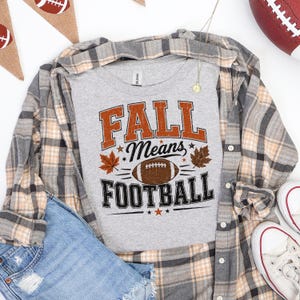 Może przedstawiać: Szary melanżowy t-shirt z napisem FALL Means FOOTBALL w kolorze pomarańczowym i czarnym, uzupełniony grafiką brązowej piłki do futbolu amerykańskiego oraz ilustracjami jesiennych liści. Koszulka jest założona pod koszulę w kratę i dżinsowe szorty.