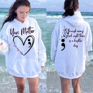 May include: A white hooded sweatshirt featuring black text and graphics. The front displays the words You Matter above a heart outline with a semicolon and the text Your story isn't over. The back shows butterflies, a semicolon, and the text Through every dark night there is a brighter day.