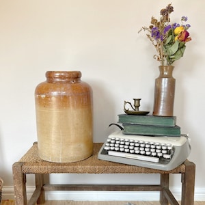 May include: A wooden bench with a woven seat, a large brown ceramic jar, a vintage typewriter, a stack of books, a brass candlestick, and a vase with dried flowers.