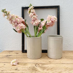 May include: Two beige ceramic vases with vertical lines, one filled with pink flowers. A black framed rectangle is behind the vases. The vases are on a wooden surface.