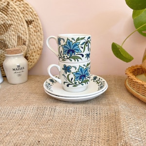 May include: Two white ceramic coffee mugs with blue and green floral designs stacked on top of each other. The mugs are sitting on a white saucer with a blue and green floral design.
