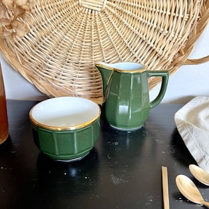 May include: A green ceramic creamer and sugar bowl set with gold trim. The creamer has a handle and a spout. The sugar bowl is round. The set is on a black surface.