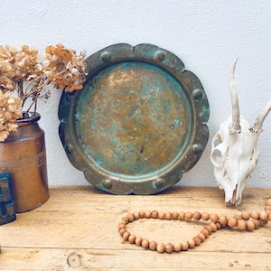 May include: A round, scalloped-edge brass tray with a weathered, oxidised patina featuring patches of green and brown. The tray is positioned between a brown ceramic jar filled with dried flowers and a white animal skull with antlers.