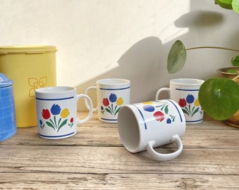 May include: Set of four white ceramic mugs with a blue, red, yellow, and green floral design. The mugs have a simple, classic shape and are perfect for everyday use.
