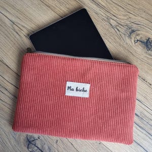 May include: A rectangular, rust-coloured corduroy zippered pouch featuring a white fabric label centred on the front with the black text Ma biche. A black tablet is partially inserted into the top opening of the pouch, revealing its screen.
