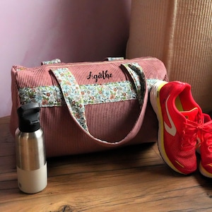 Peut inclure: Un sac de sport en velours côtelé rose poudré avec des sangles à motifs floraux et le nom "Agathe" en écriture noire. Une gourde en acier inoxydable et des chaussures de course rouges avec des accents blancs et jaunes sont à côté du sac. Sur une surface en bois.