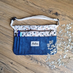 Peut inclure: Un sac banane en velours côtelé bleu avec un panneau en tissu blanc à motif léopard marron et noir. Le sac comprend un compartiment zippé, deux emplacements pour stylos, une sangle blanche réglable et une étiquette blanche avec le nom Katia.