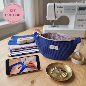 Peut inclure: Un sac banane en velours côtelé bleu avec une étiquette blanche marquée M-P, accompagné d'un kit de couture comprenant des tissus, une fermeture éclair, une sangle blanche, une coupelle dorée avec des épingles et un smartphone.