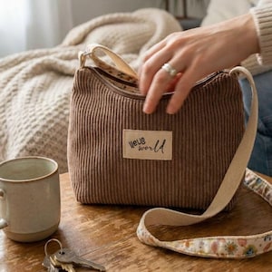 Op de afbeelding: Een bruine schoudertas van corduroy met een rechthoekige beige stoffen label met de tekst "hello world" in zwarte schrijfletters. De tas heeft een lange, verstelbare schouderband met een bloemenpatroon aan de onderkant en een ritssluiting.