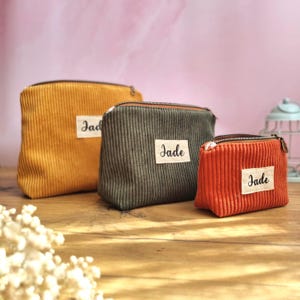 May include: Three corduroy zip-up pouches in mustard yellow, olive green, and burnt orange. Each pouch features a rectangular white fabric label sewn on the front with the name Jade printed in black script. The pouches vary in size.