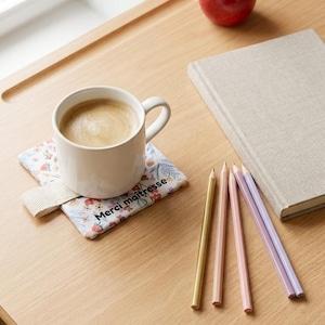 Può includere: Una tazza di ceramica bianca piena di caffè è appoggiata su un sottobicchiere floreale con la scritta "Merci maîtresse". Accanto ci sono matite colorate e un libro chiuso. Una mela rossa è appoggiata sullo sfondo su una superficie di legno.