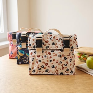 Pode incluir: Três lancheiras retangulares com alças superiores e fechos de fivela de plástico preto. A bolsa frontal tem um fundo bege com manchas de leopardo castanhas e pretas. A bolsa do meio é azul-escura com padrões de folhas douradas e a de trás é rosa.